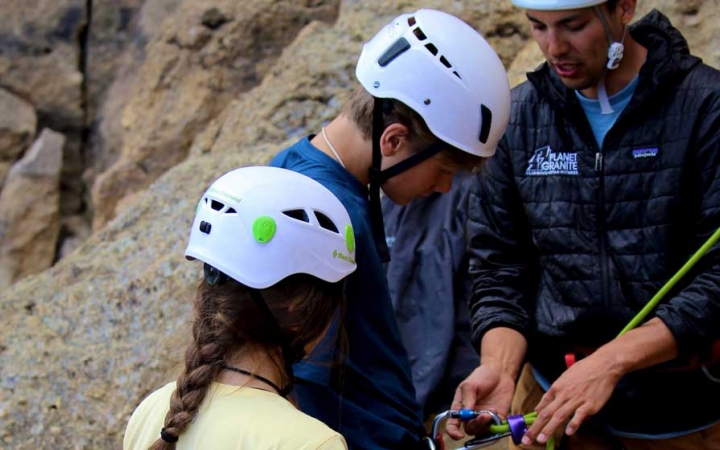 An instructor helps to tie a knot and secure it to a student's harness, while another student looks on.